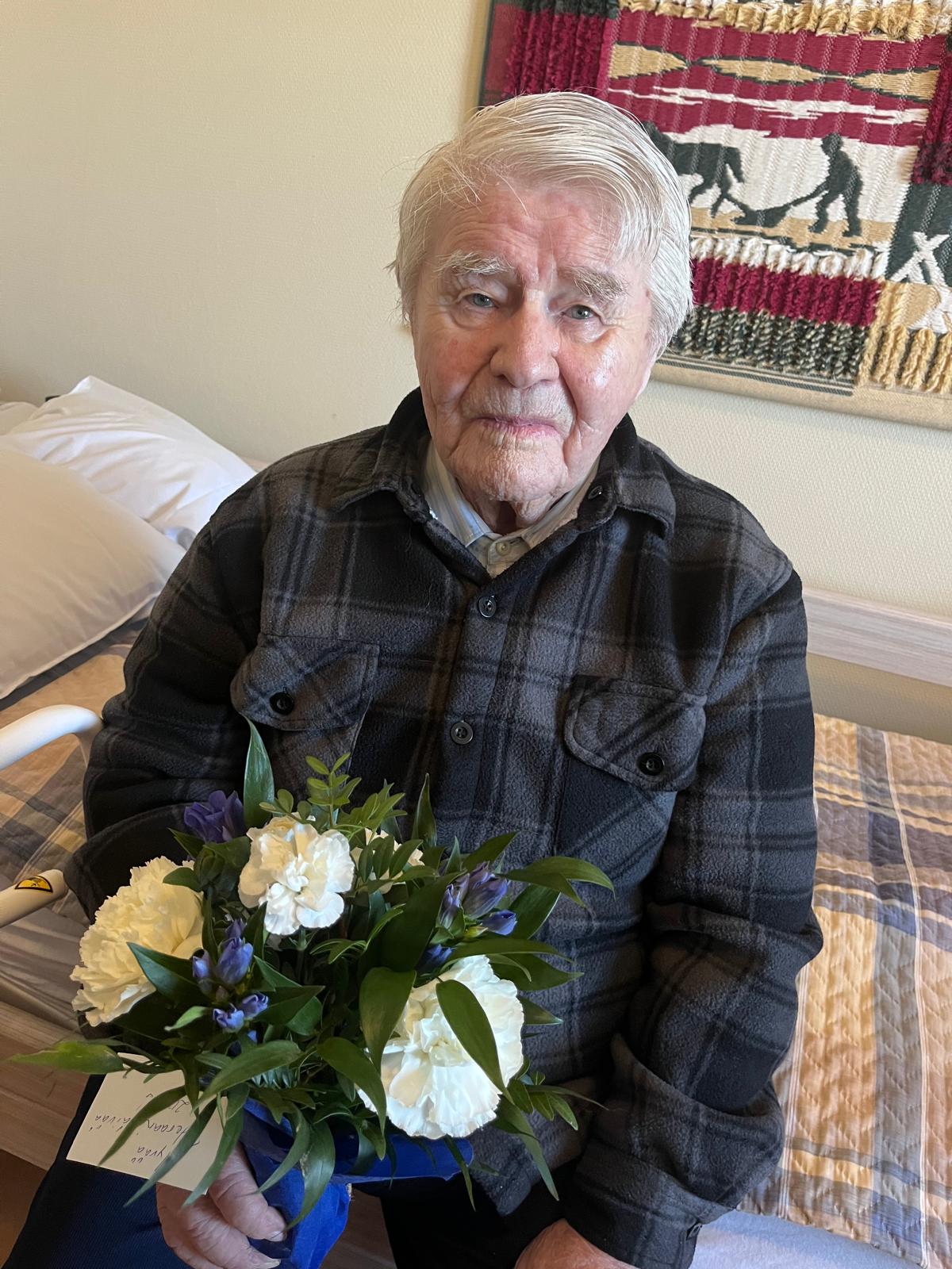 Veteraani Jouko istuu sängyn reunalla ja pitelee kukkakimppua kädessään.