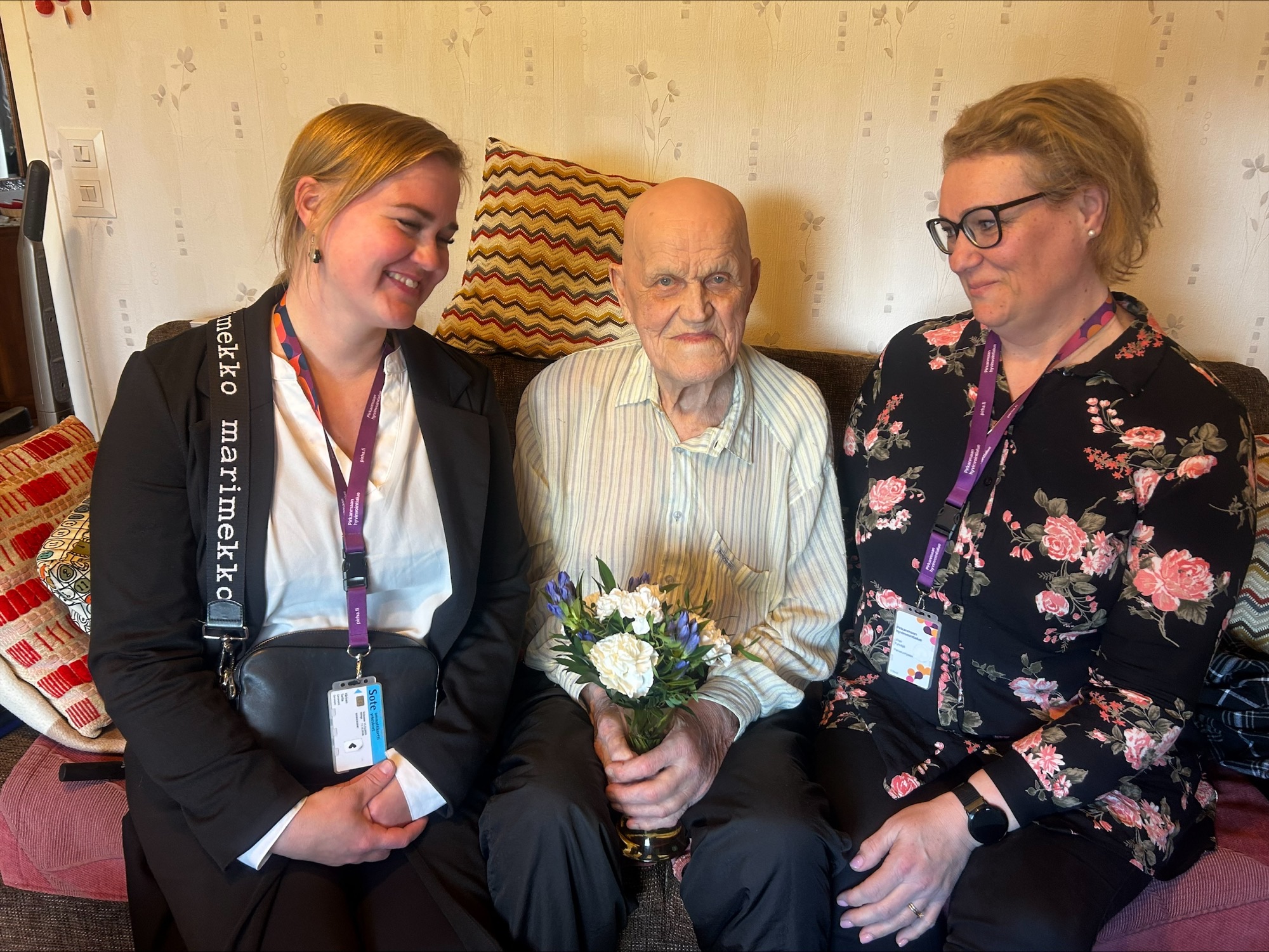 Sohvalla istuu vasemmalta oikealle: palveluohjaajat Sarita Värinen, Voitto kukkakimppu käsissä ja palveluohjaaja Virpi Kylväjä. 