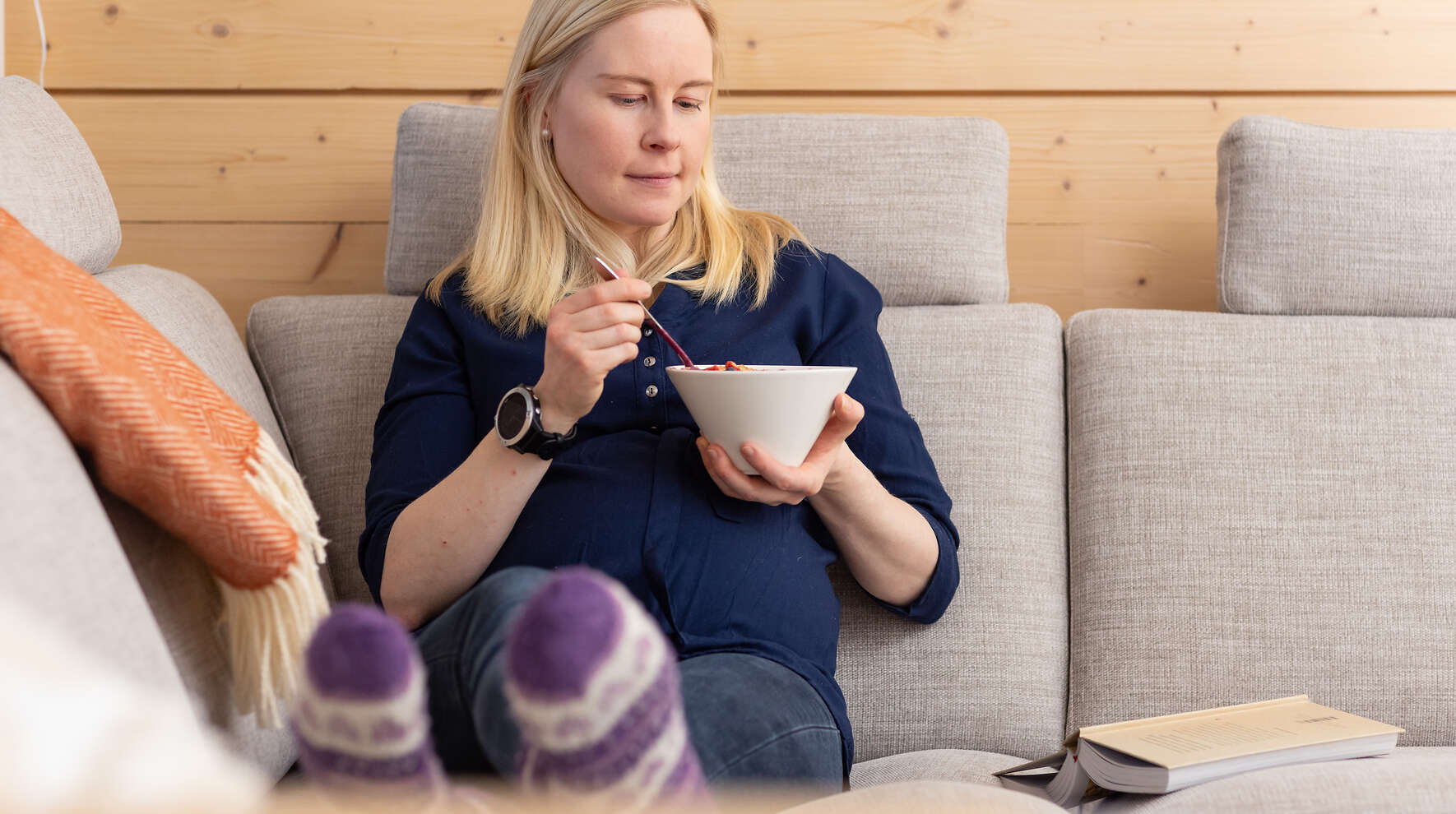 Raskaana oleva nainen istuu sohvalla ja syö jogurttikupista.