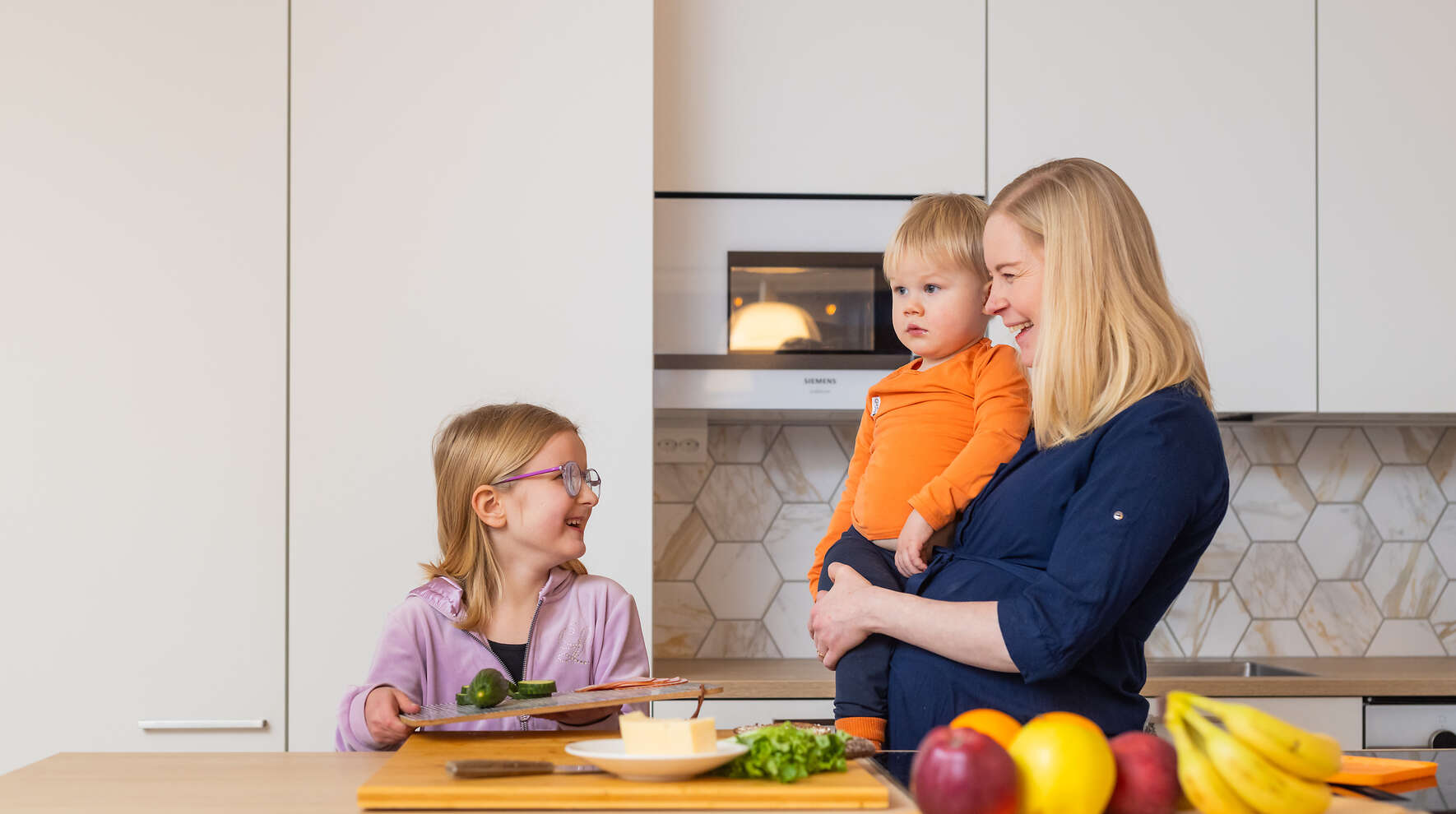 Keittiösaarekkeen takana alakouikäinen tyttö tekemässä välipalaa. Raskaana oleva nainen seisoo vieressä pienempi lapsi sylissään ja hymyilee tytölle.