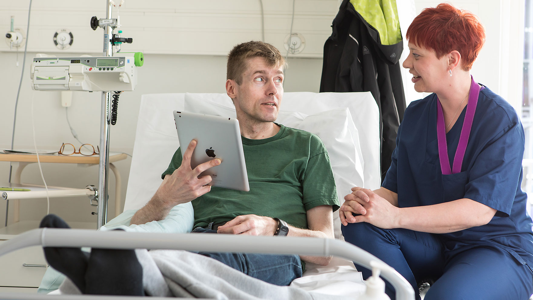 Miespotilas juttelee sairaalasängyllä vieressään istuvan hoitajan kanssa.